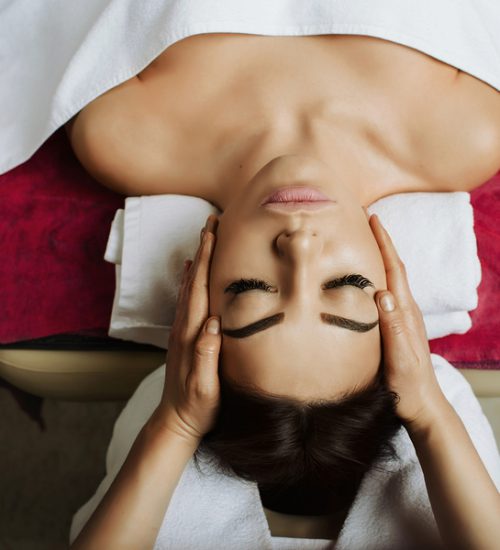 Attractive brunette woman getting massage spa treatment lying on a massage table, hands working on massaging woman's head. Relaxing facial massage at spa salon, top view.